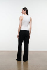 A woman stands with her back to the camera wearing a fitted sleeveless white top and black wide-leg pants in a minimalist studio with a white wall and beige floor.