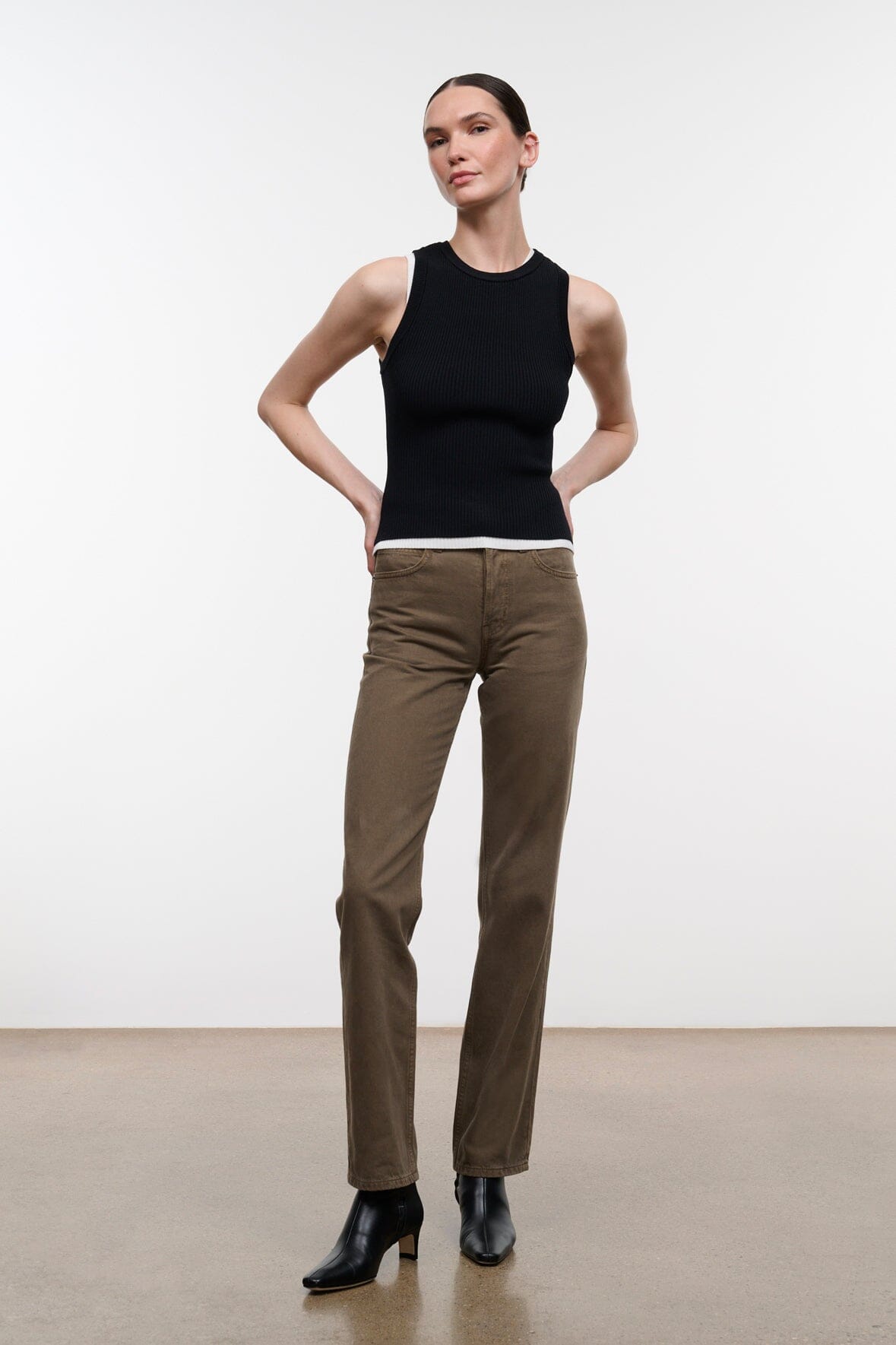 A model stands in a studio wearing a black sleeveless top, brown straight-leg pants, and black ankle boots against a plain light background.