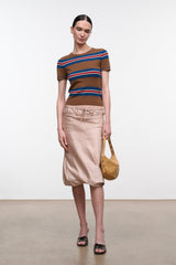 A model stands in a studio wearing a brown short-sleeve knit top with blue and red horizontal stripes, paired with a light beige midi skirt and holding a tan shoulder bag.