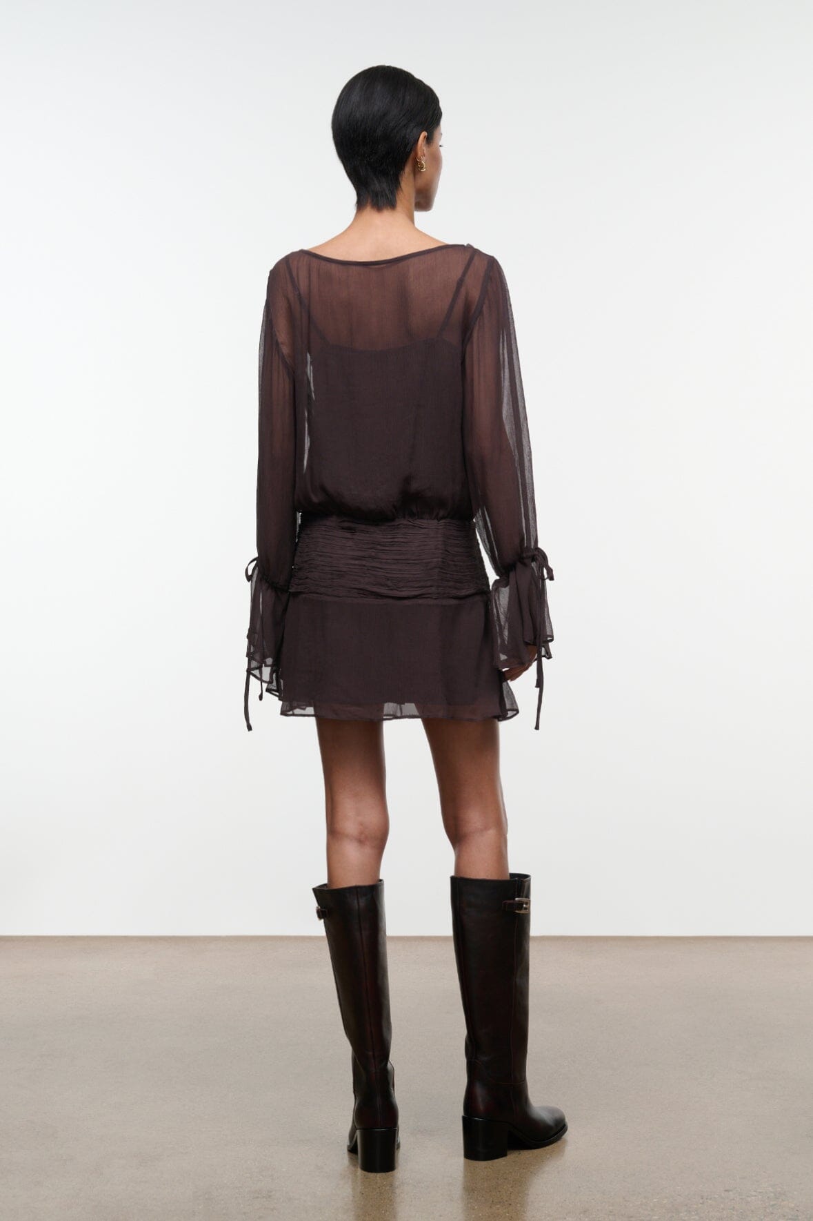 Dark brown sheer long-sleeve mini dress with ruched waist and tie-cuff details, back view.