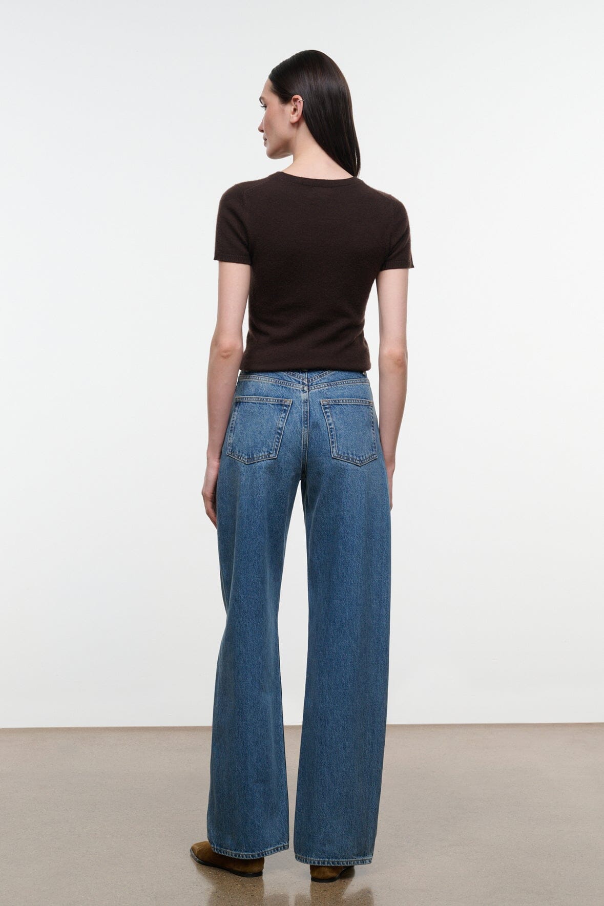 A woman shown from the back wearing a dark brown short-sleeve top and blue wide-leg jeans, standing against a plain white studio wall.