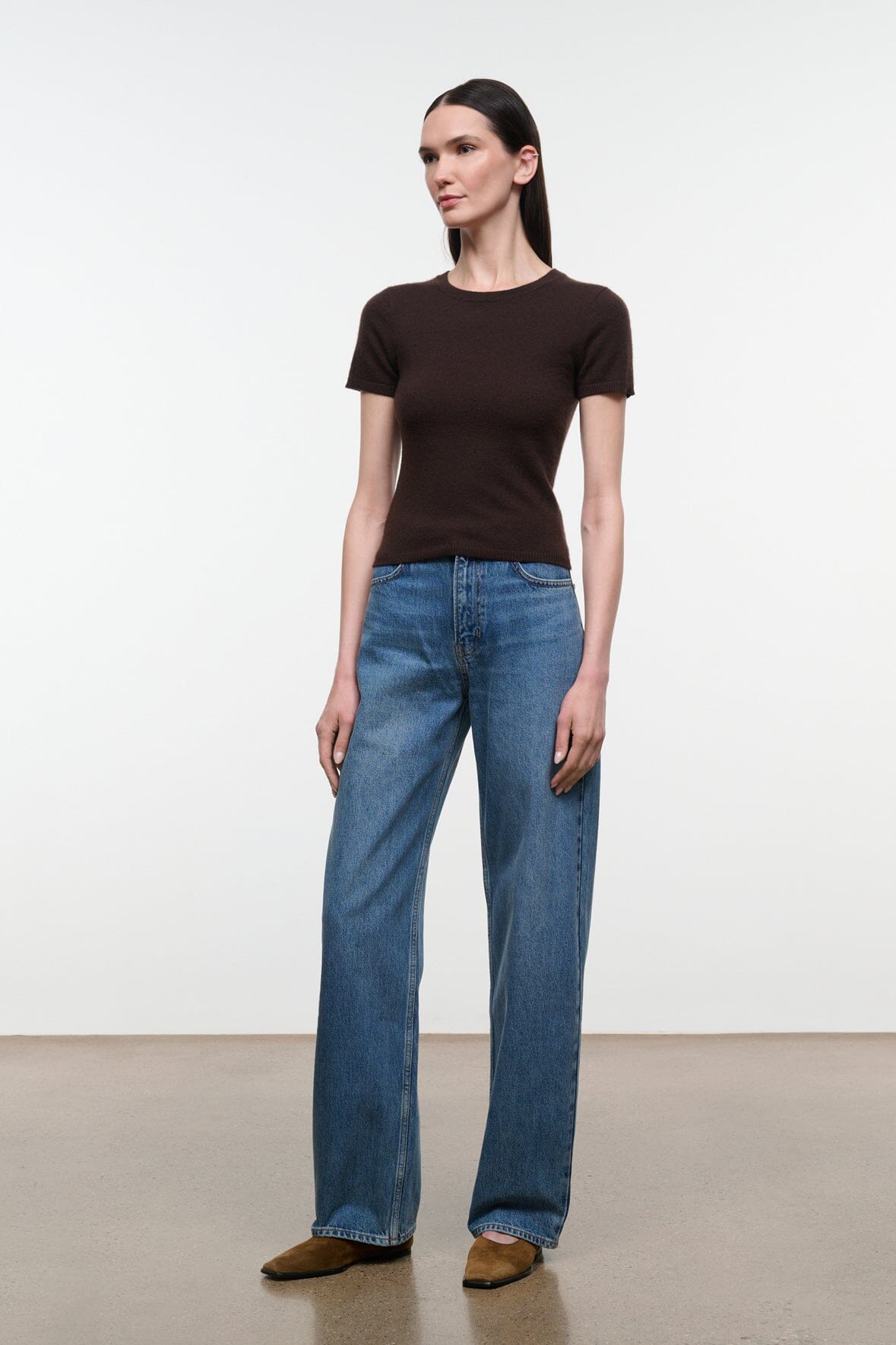 A woman modeling a dark brown short-sleeve crewneck t-shirt and wide-leg blue jeans, standing against a plain white studio background.