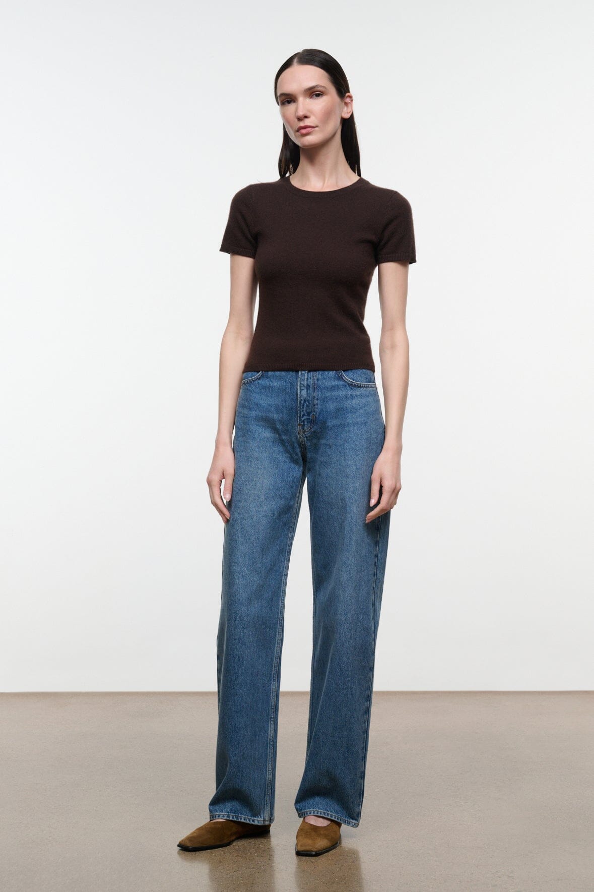 A woman modeling a dark brown short-sleeve crewneck t-shirt and wide-leg blue jeans, standing against a plain white studio background.
