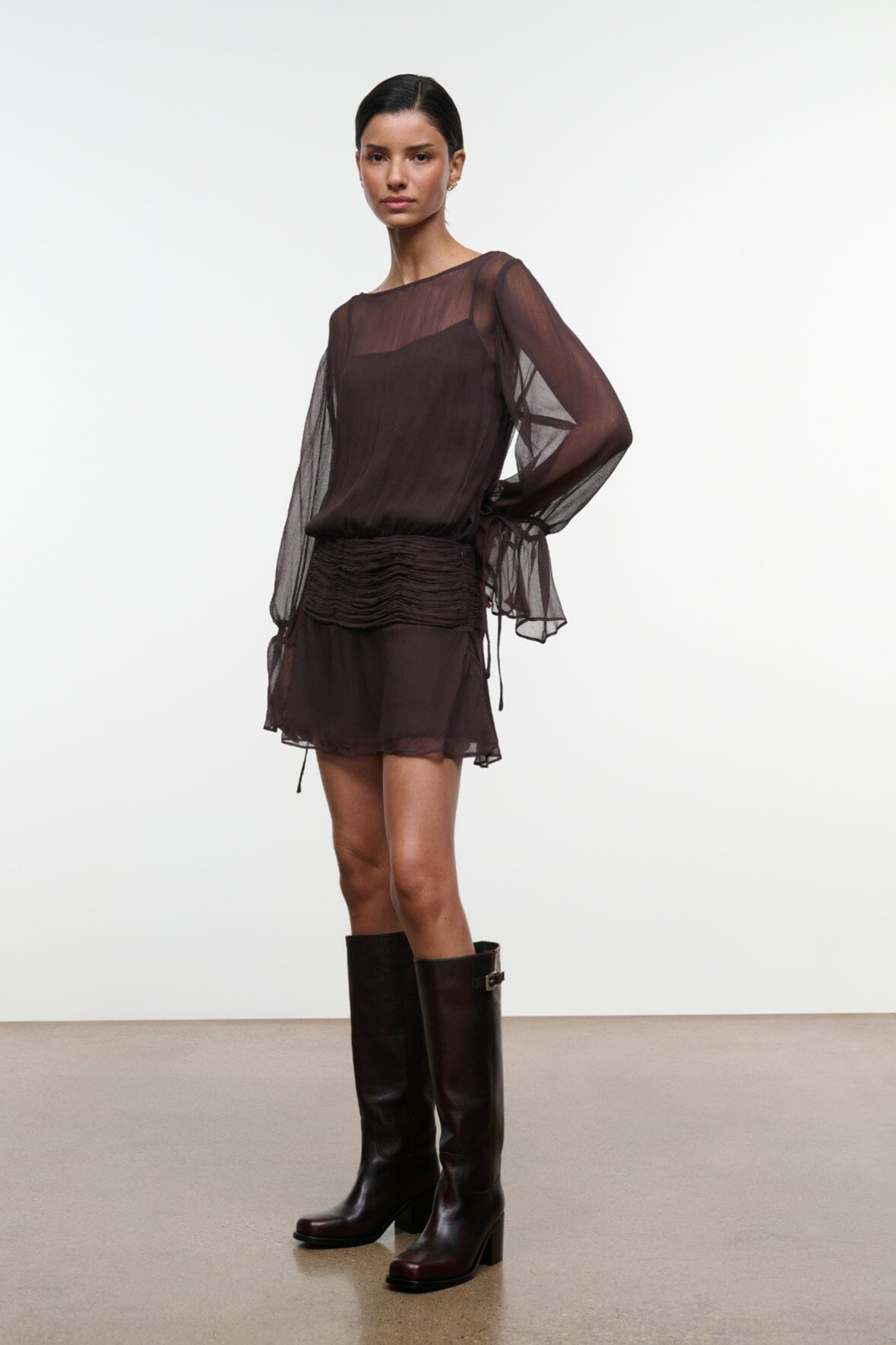 Dark brown sheer long-sleeve mini dress with ruched waist and draped tie cuffs, side view.
