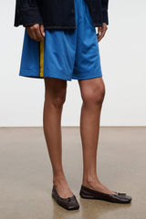 Espresso brown ballet flats with bow detail and stitched trim, worn with blue shorts.
