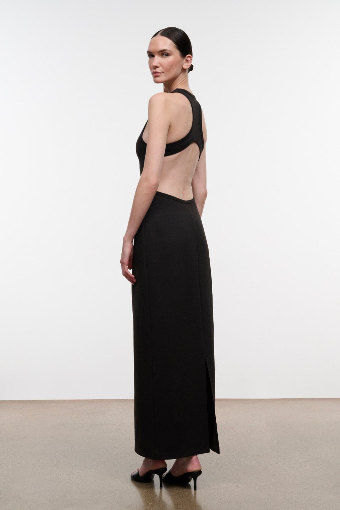 A woman modeling a floor-length black dress with a dramatic open back and halter-style shoulder straps, shown from the back in a studio setting.