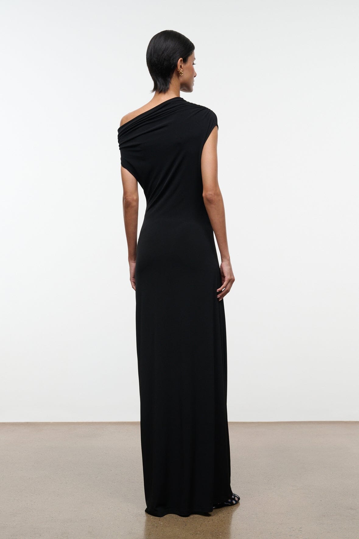 Black one-shoulder draped maxi dress, sleeveless, back view with floor-length skirt