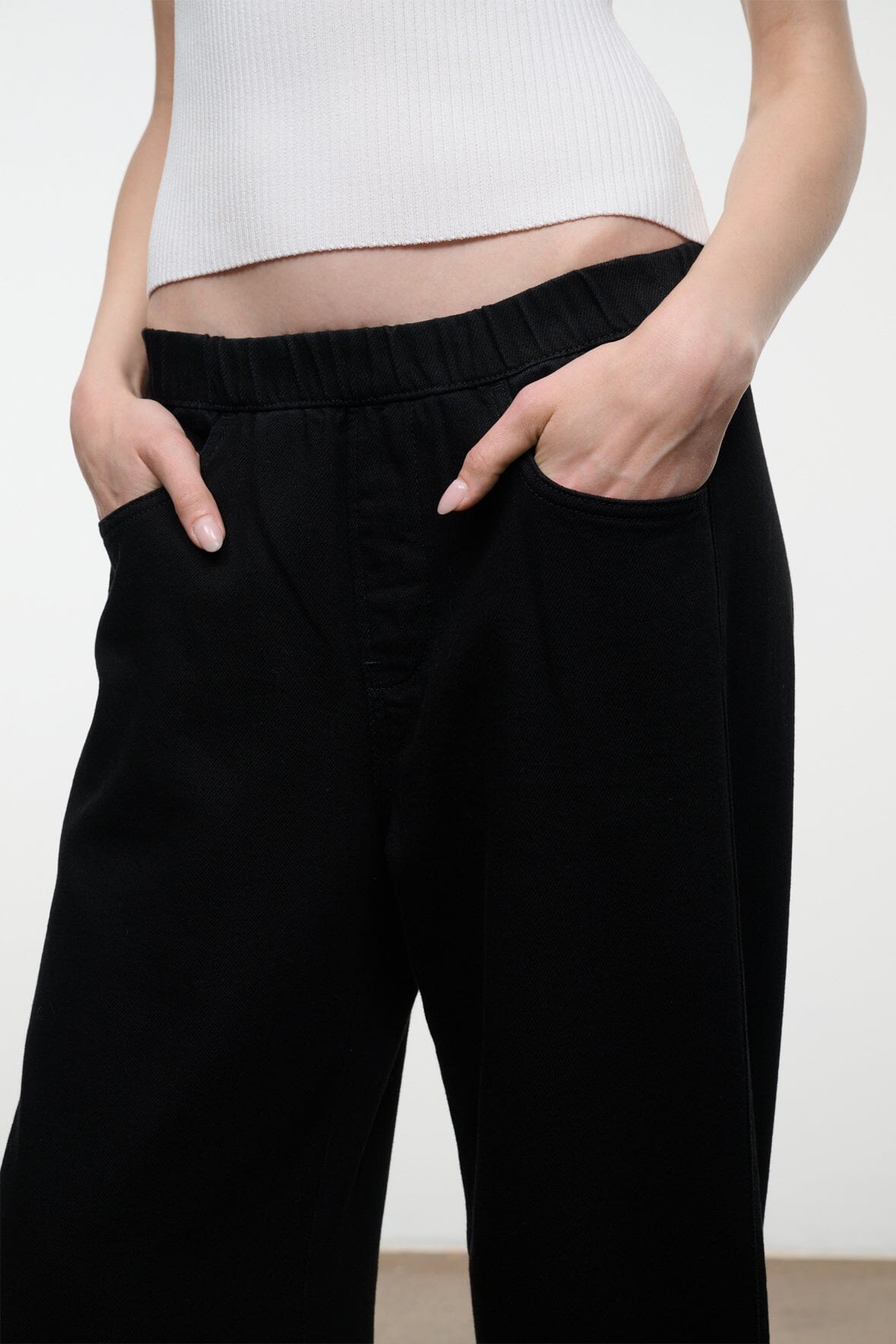 A close-up of a person wearing black wide-leg pants with an elastic waistband and side pockets, paired with a white ribbed crop top against a plain light background.