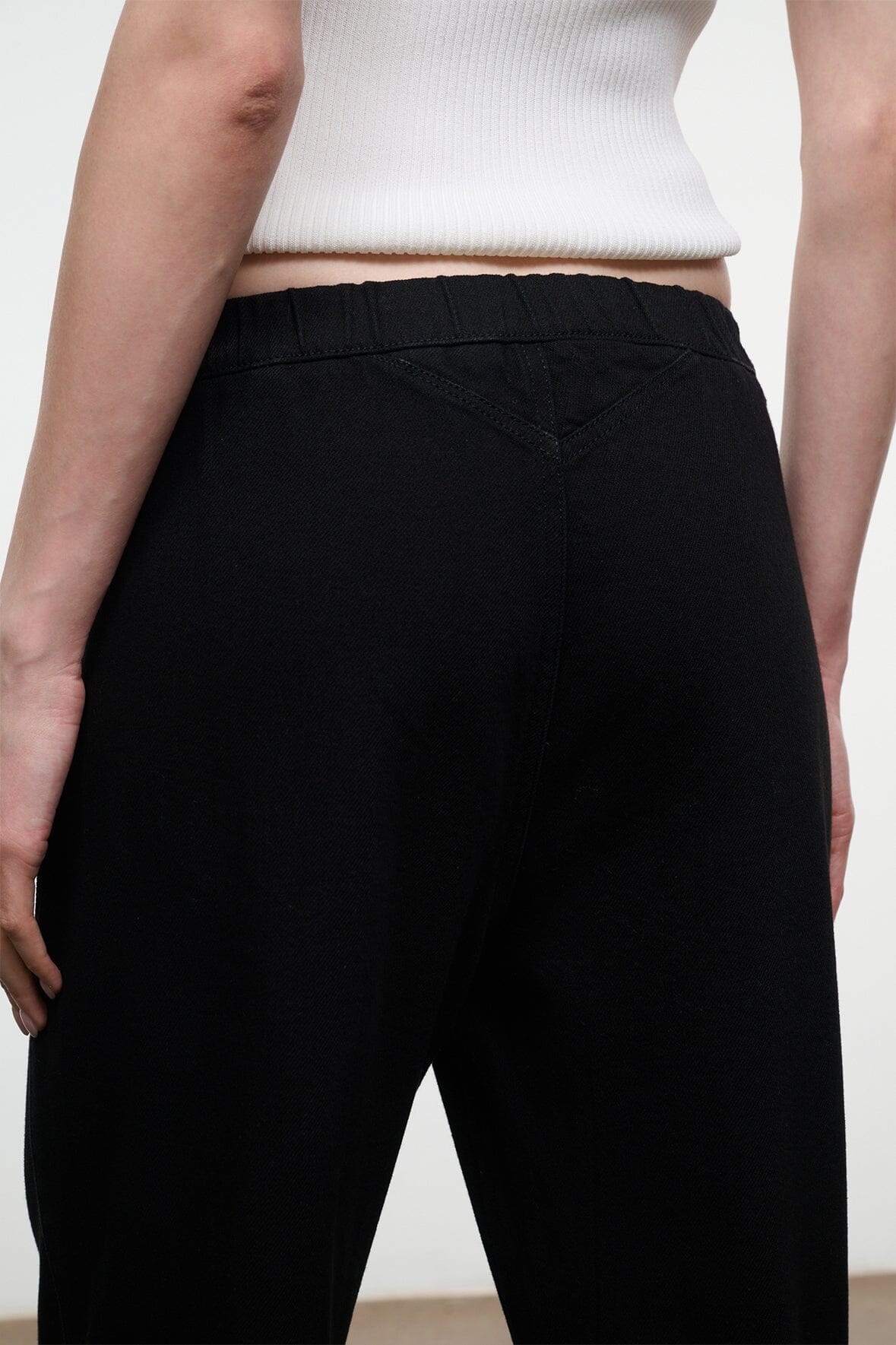 Close-up back view of a person wearing black wide-leg trousers and a white ribbed crop top against a plain light background.