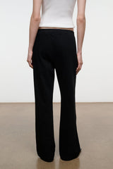 A model shown from the back wearing black wide-leg trousers and a white ribbed top, standing against a minimalist studio background with a white wall and beige floor.