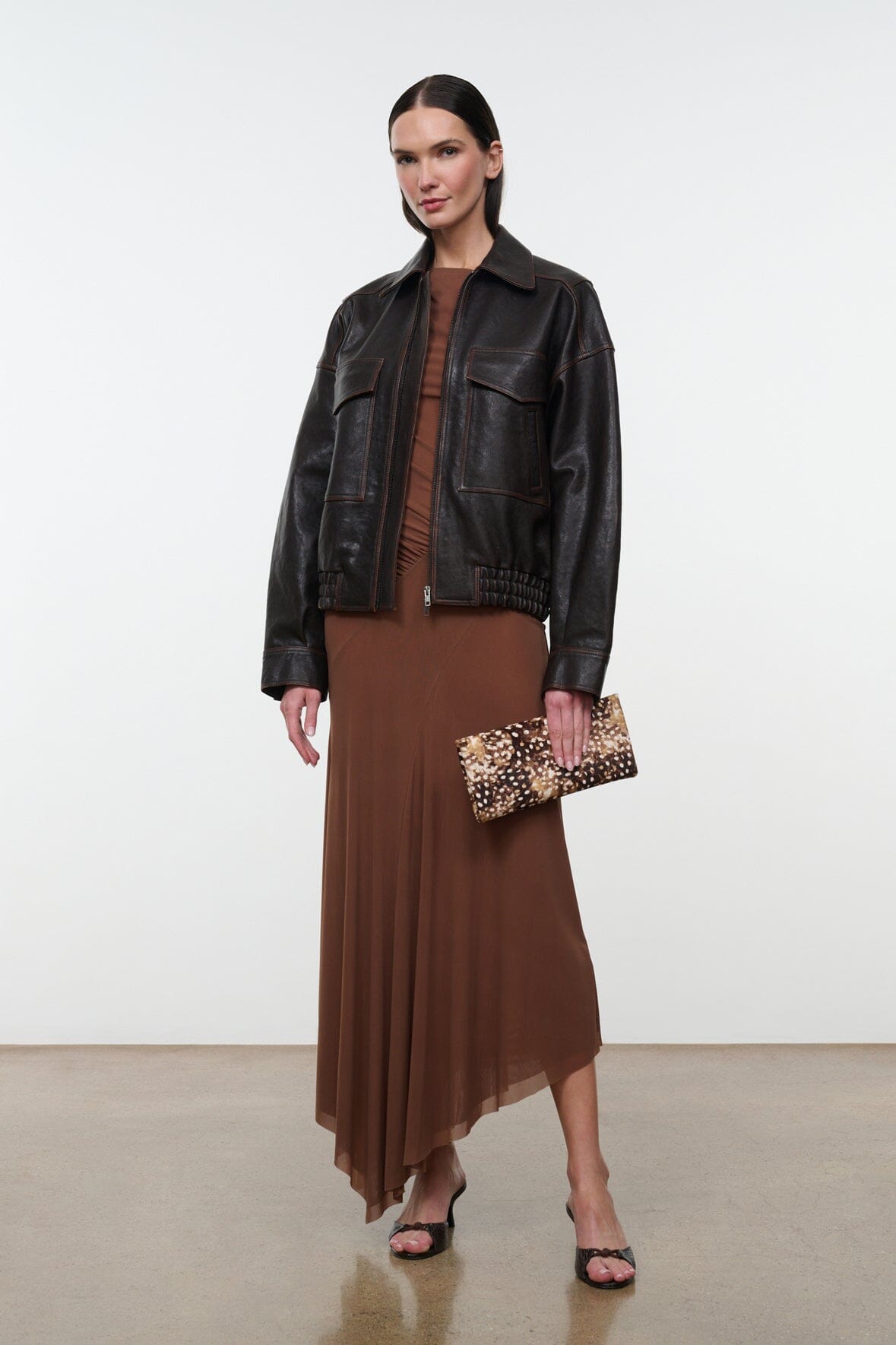 Brown one-shoulder ruched dress under a black leather jacket; asym hem visible