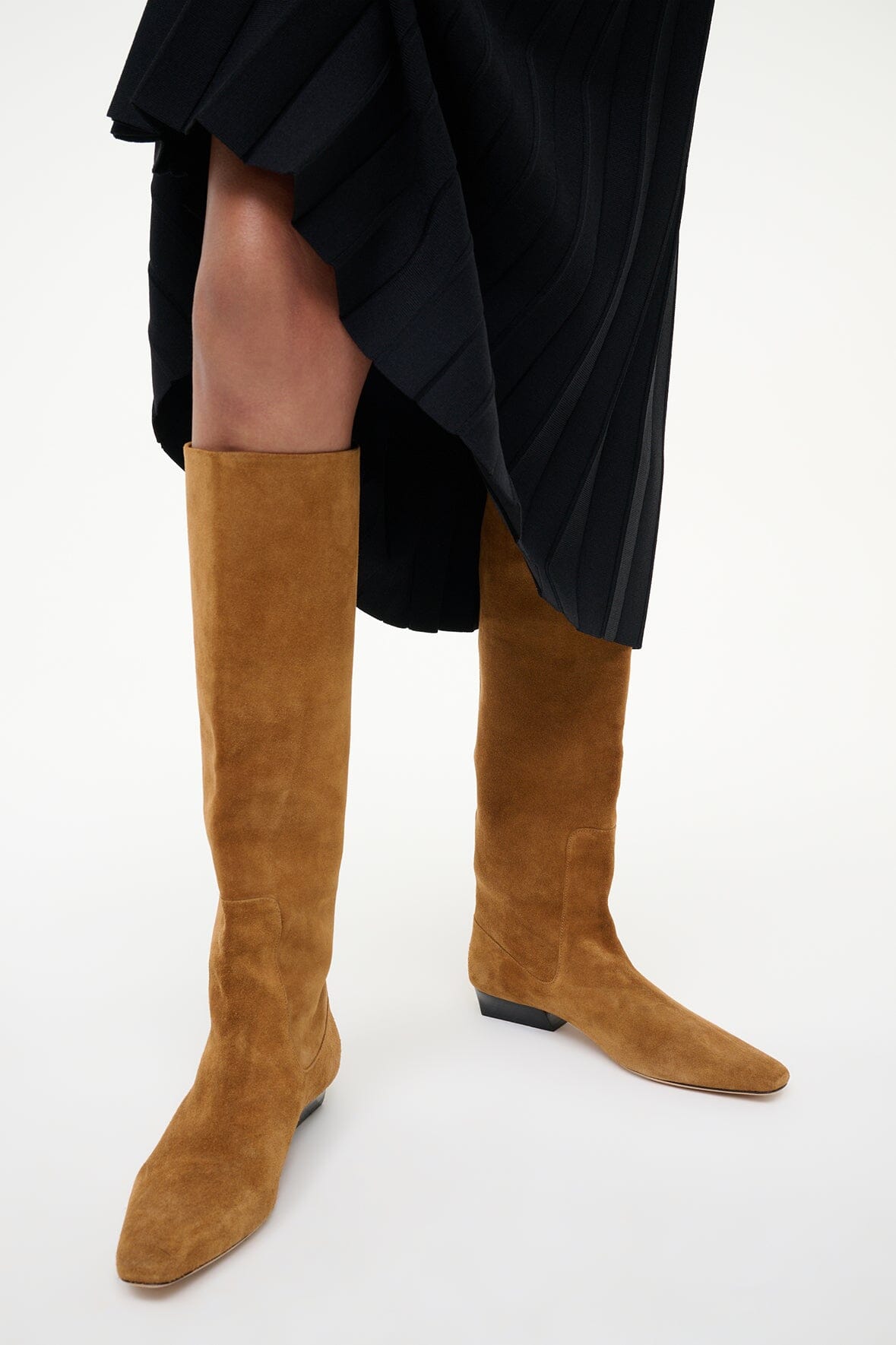Tan suede flat knee-high boots with low stacked heel under a black dress hem