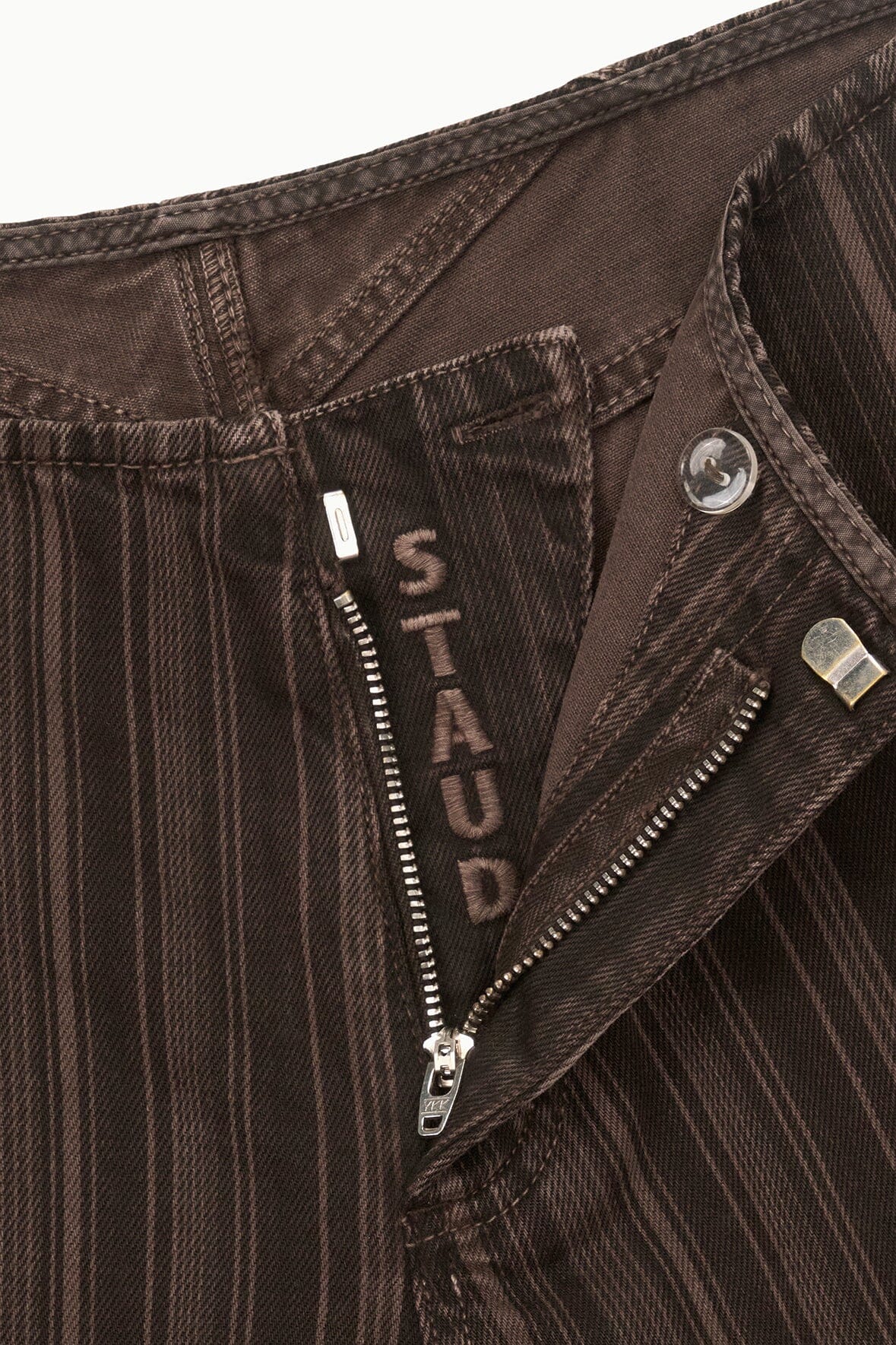 Dark brown pinstripe jeans close-up with zip fly, button, and logo patch