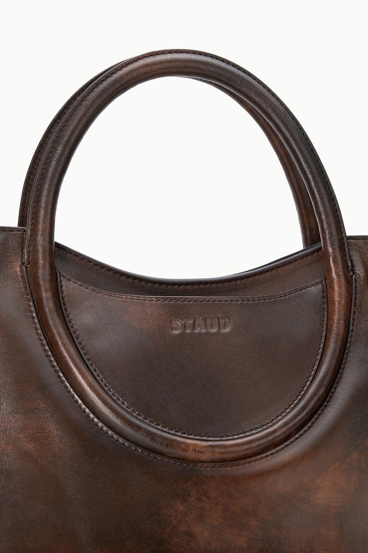 Dark brown distressed leather satchel close-up: oval handle edge and 'STAUD' logo