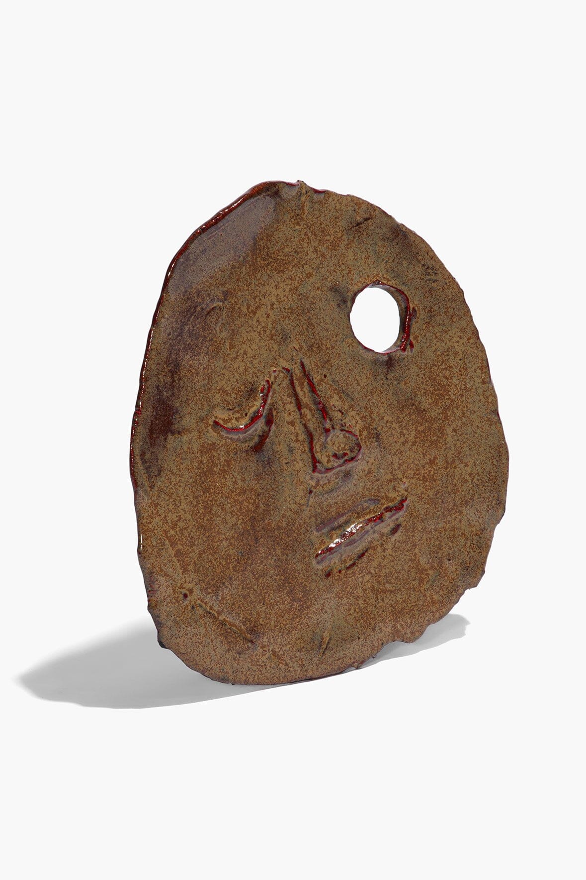 Ceramic face ornament in molasses brown glaze, round cutout hole, sculpted nose and mouth.