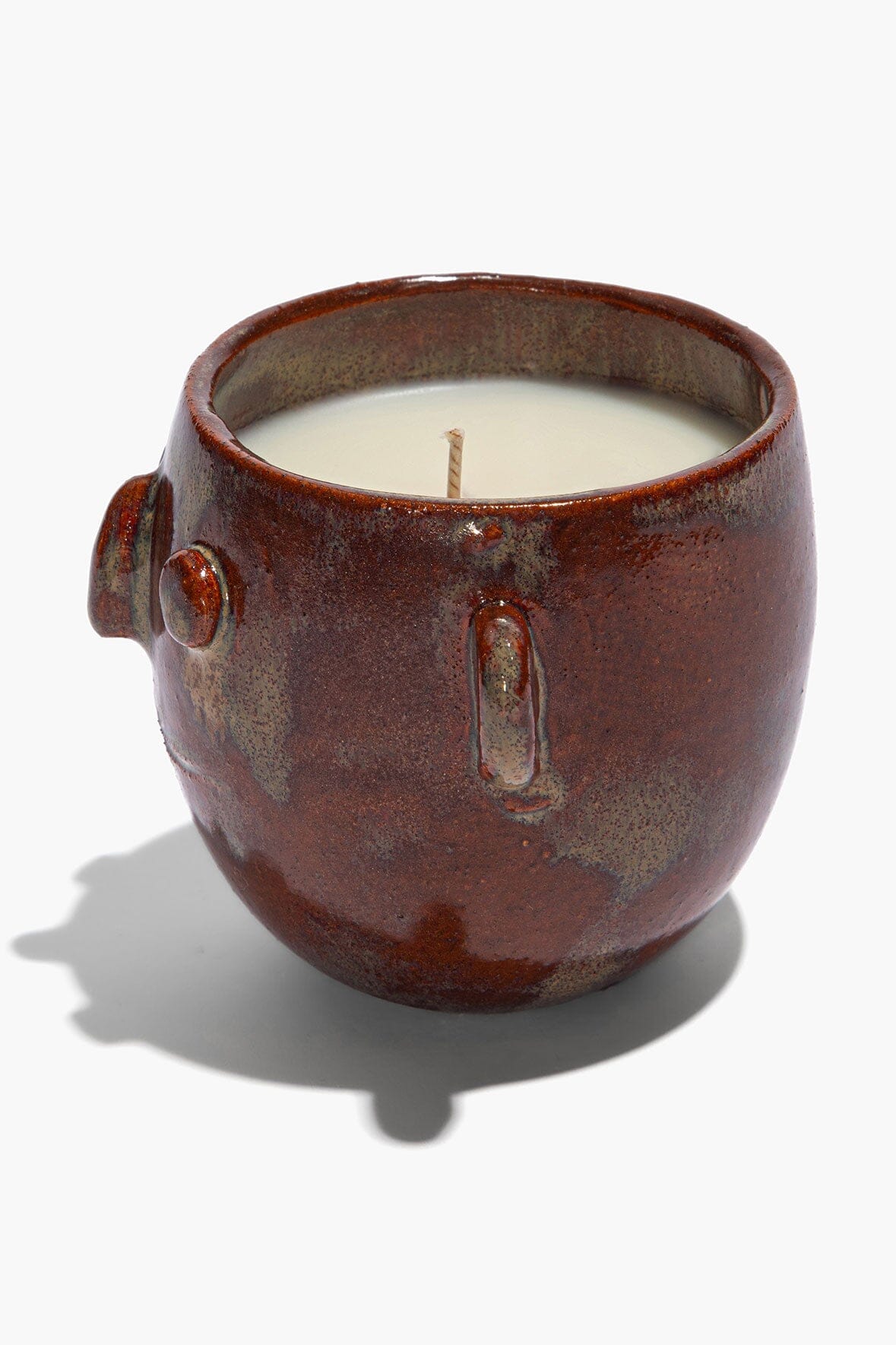 Ceramic candle pot with vertical face emblem and curved rim; white wax with wick