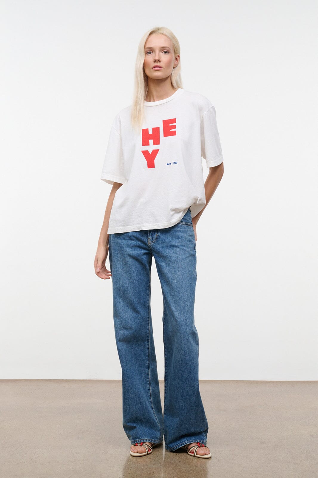 White relaxed tee with red “HEY” logo; worn with blue jeans, hem slightly draped.