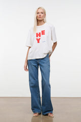 White relaxed tee with red “HEY” logo; worn with blue jeans, hem slightly draped.