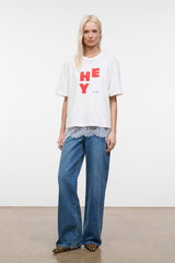 White relaxed tee with red “HEY” graphic; worn with blue jeans, slightly oversized fit.