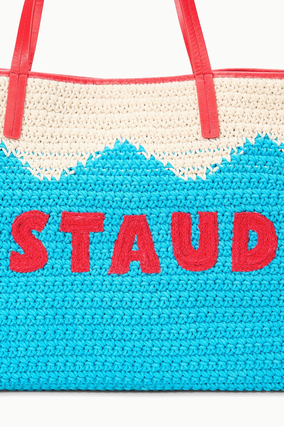 Blue knit tote bag with cream upper section and red handles, “STAUD” lettering in red